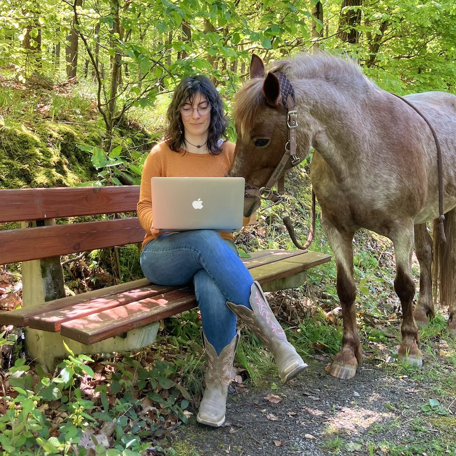 Texterin Mirjam mit ihrer Ponystute sitzt auf einer Bank mit Laptop